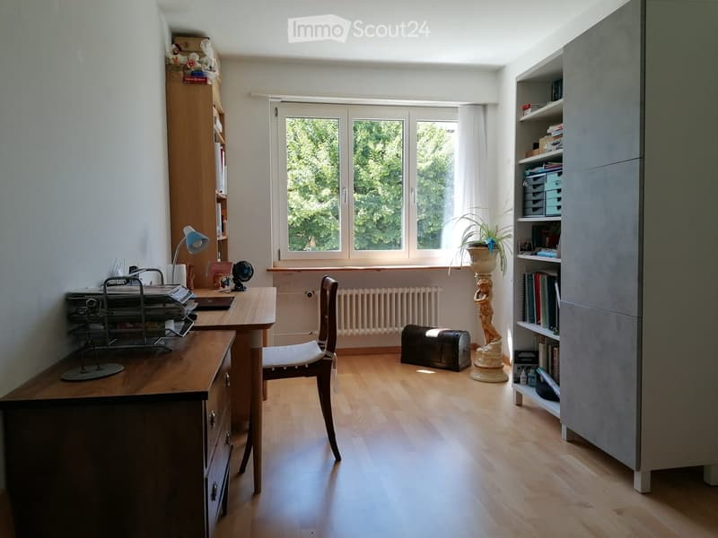 Brightly lit room with wooden flooring, large window, desk with chair, bookshelf, cabinet, and decorative items including a plant and a statue.