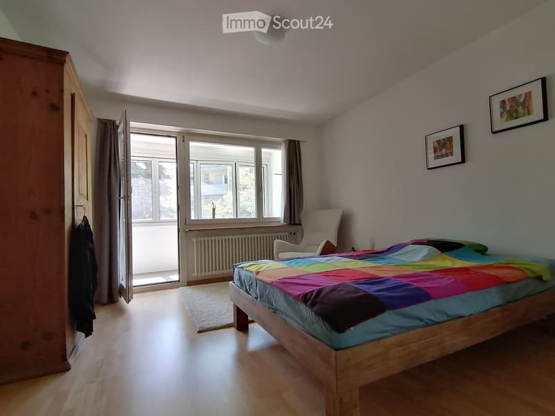 Bright bedroom with wooden flooring, large windows, a colorful bedspread, a single chair, and a radiator under the windows.