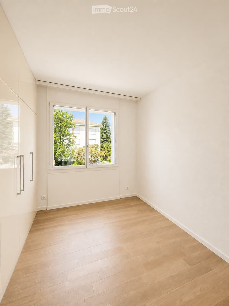Empty room with light wooden flooring, white walls, and large windows offering a view of greenery outside.