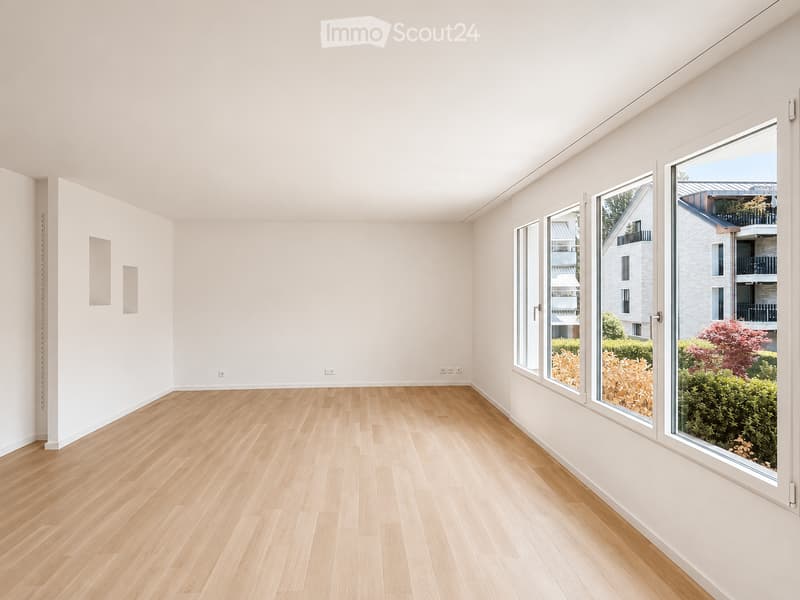 Empty room with light wooden flooring and white walls, featuring large windows with a view of greenery and neighboring buildings.