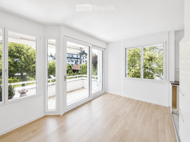 Bright, unfurnished room with light wooden flooring, white walls, large windows, and sliding glass doors leading to a balcony.