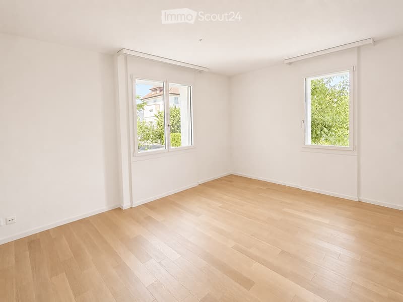 Empty room with light wooden flooring and white walls, featuring two windows with white frames and blinds, and a visible electrical outlet on the left wall.