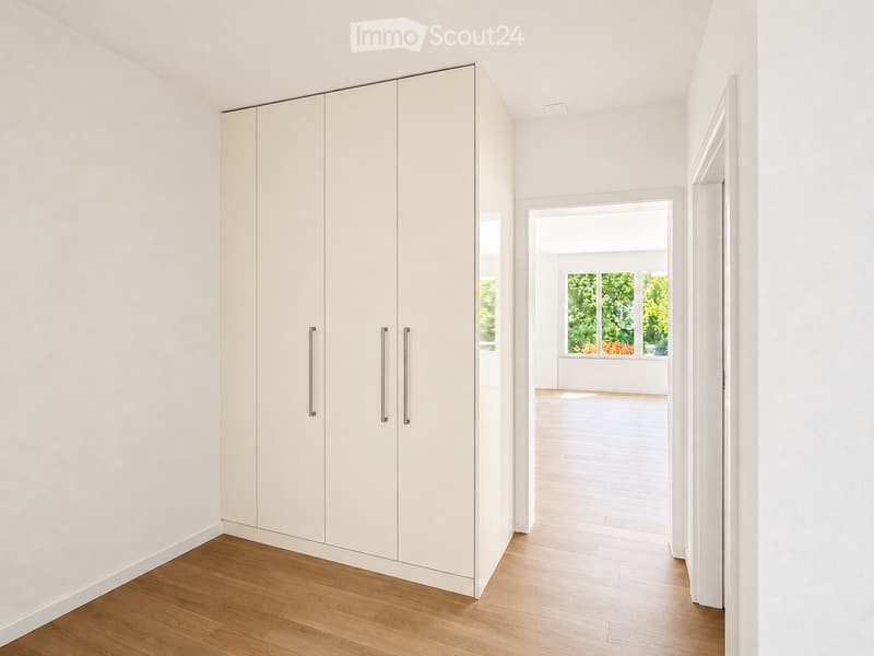A clean, modern hallway with light-colored wooden flooring, white walls, and built-in storage units with sleek handles. The hallway leads to a bright, open space with large windows and natural light.
