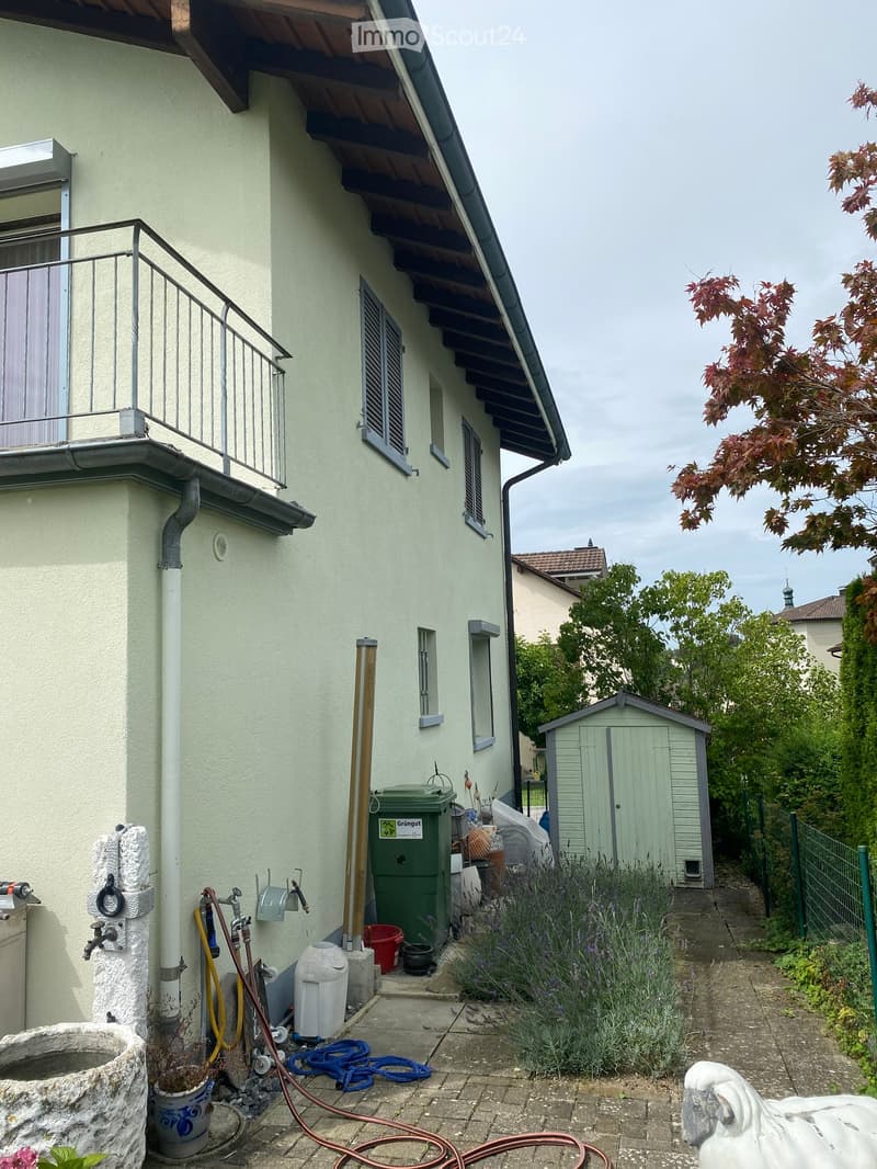 Garden area adjacent to a house with a balcony, featuring a small shed, plants, a water fountain, and various garden tools and equipment.