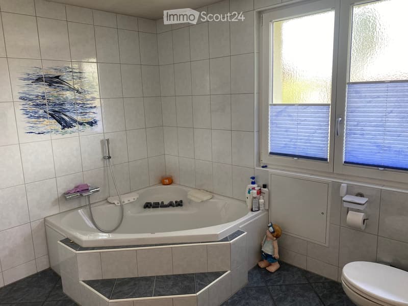 Modern bathroom with a large bathtub, toilet, and a window with blinds. The bathtub features a decorative dolphin mural on the wall and a small toy figure nearby.
