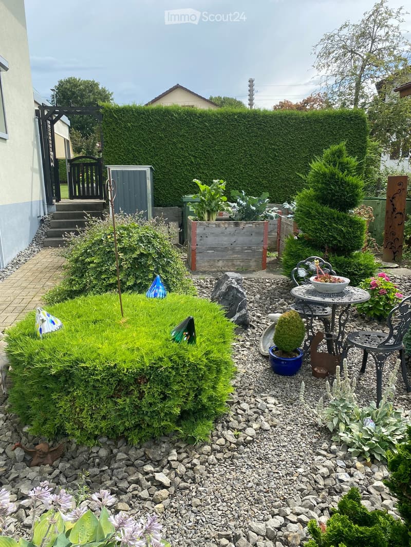 Well-maintained garden with a variety of plants, shrubs, a small seating area with a table and chairs, and a wooden planter box. The garden features a gravel pathway, a stone border, and a neatly trimmed hedge as a backdrop.