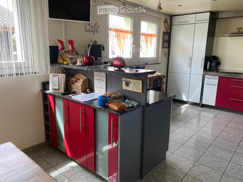 Modern kitchen with red and grey cabinets, granite countertop, and various items on the counter including a vase, cup, and handbag. There are windows with orange curtains and a sign on the wall.