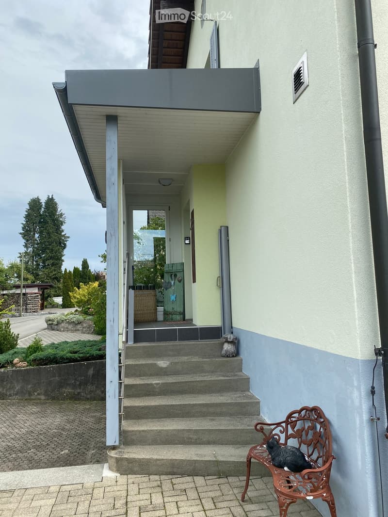 Two-story house with a covered entrance, steps leading up, and a small balcony area above. A decorative metal chair with a cat is placed near the entrance.