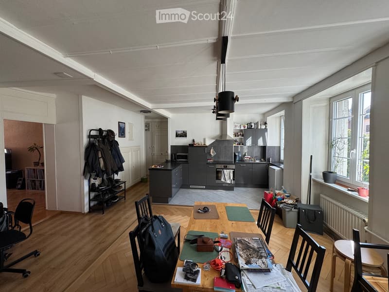 Open-plan living and dining area with a wooden dining table, chairs, a backpack, and a kitchen area with cabinets, an oven, and a refrigerator.