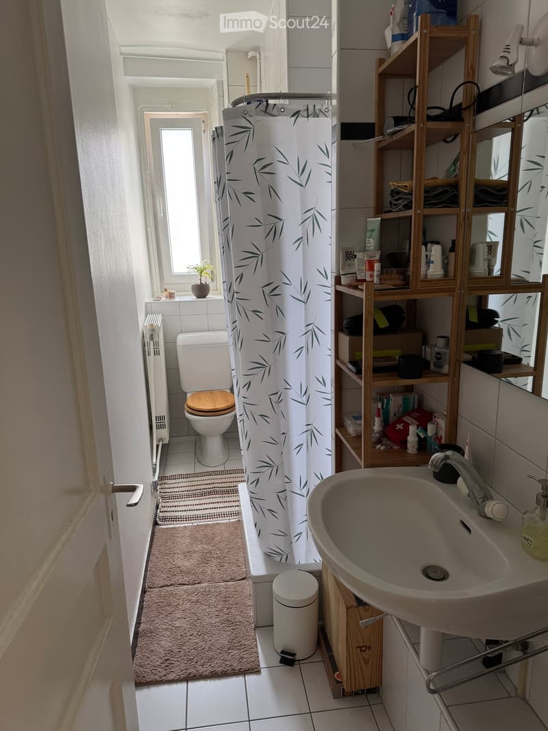 Modern bathroom with white tiles, a toilet with a wooden seat, a sink with a faucet, a shower curtain with a leaf pattern, a wooden shelf holding toiletries, a small plant on the windowsill, and a radiator.