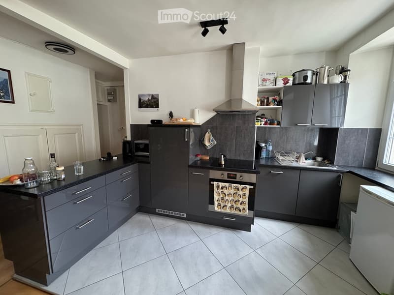 Modern kitchen with grey cabinets, black countertops, and stainless steel appliances. Features include a refrigerator, oven, sink, and various kitchen items on the counters.