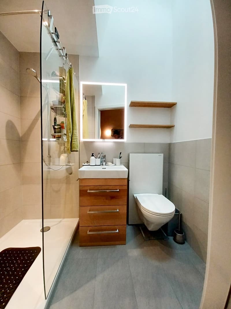 Modern bathroom with a white sink, wooden cabinet, toilet, mirror, and a glass-enclosed shower. Wooden shelves are mounted on the wall, and a green towel hangs on the shower door.