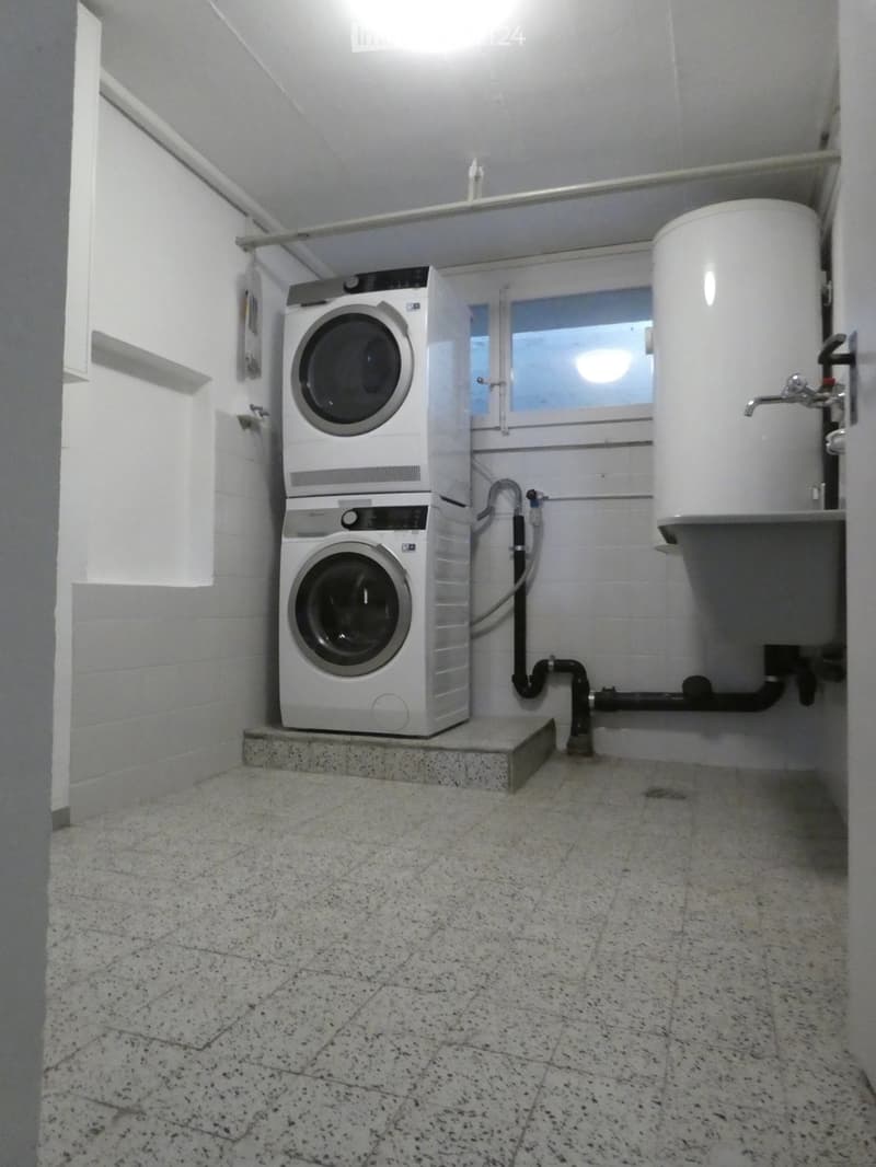A small utility room with a washing machine and a water heater, featuring white tiled walls and a speckled tiled floor.