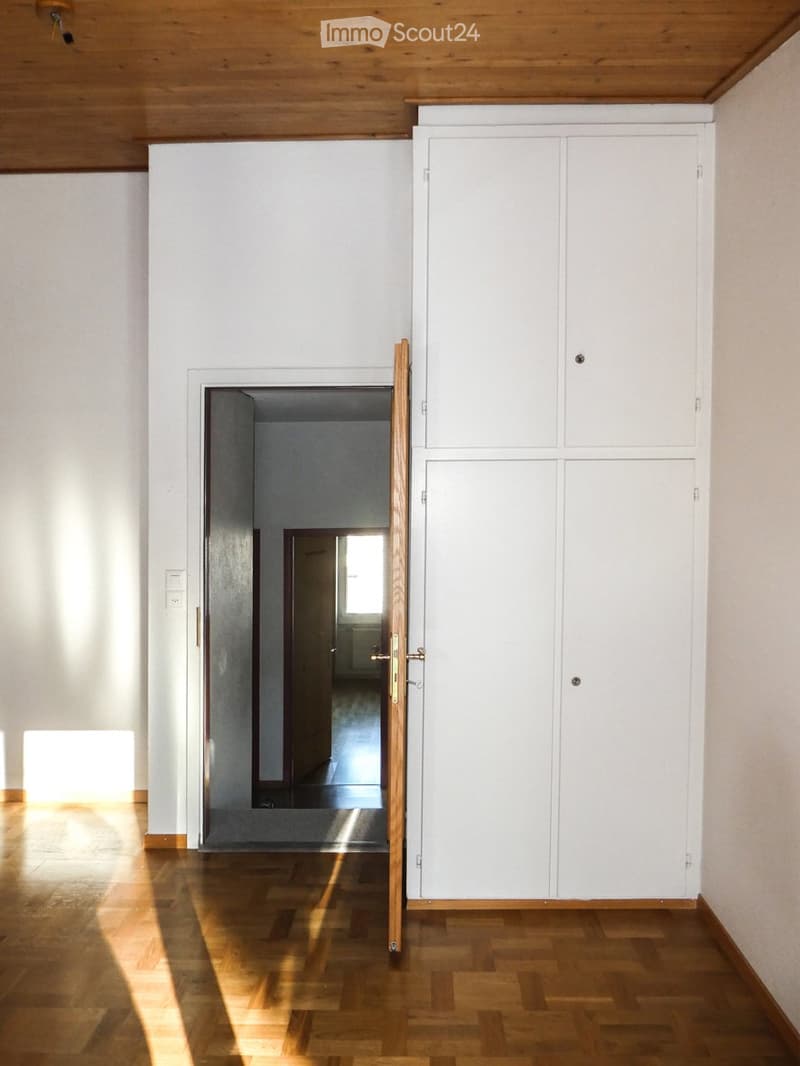 Empty room with wooden flooring, white walls, and a built-in white cabinet. Sunlight streams in from an open doorway leading to another room.