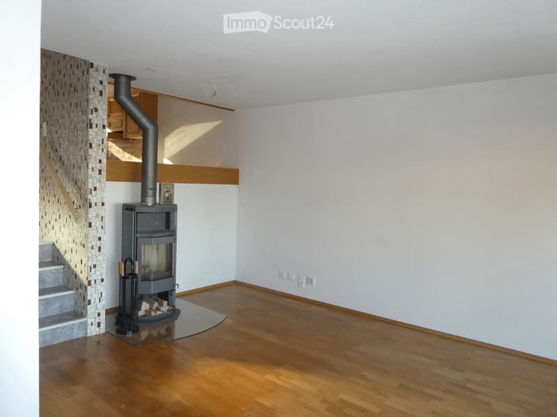 Empty room with wooden flooring, white walls, and a modern wood-burning stove with a chimney. The room has a staircase leading upwards and is well-lit with natural light.
