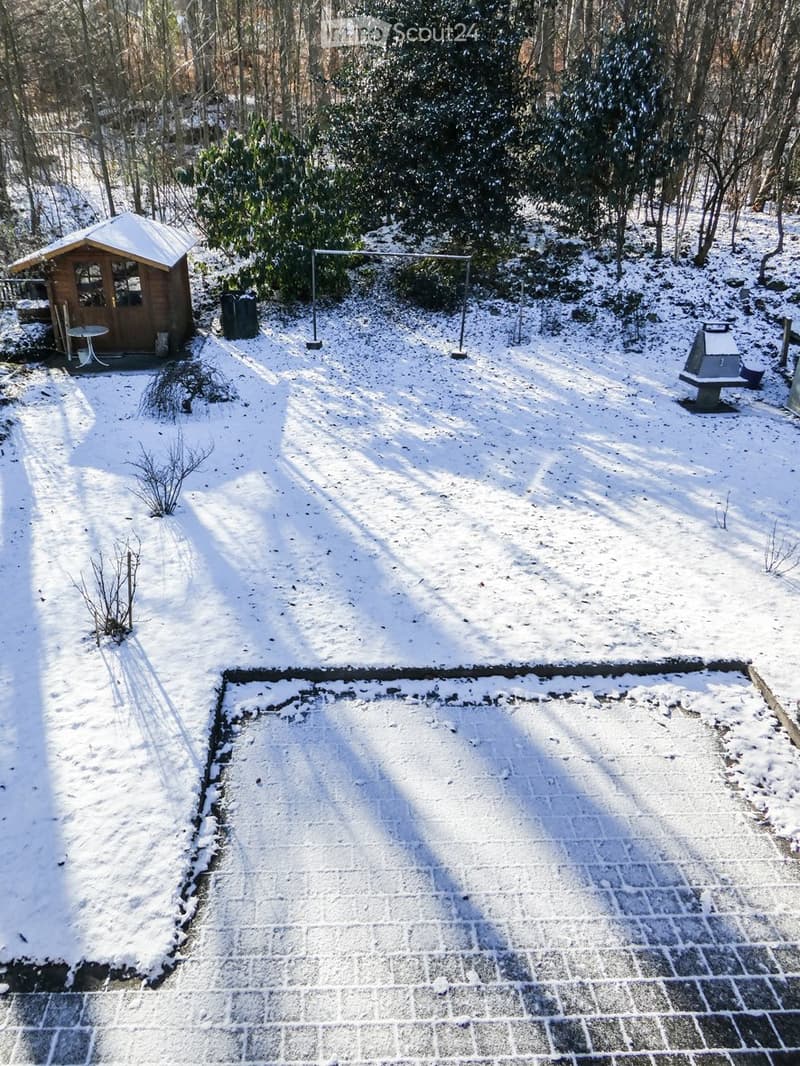 Snow-covered garden with a small wooden shed, a round table, a swing set, and a paved area.