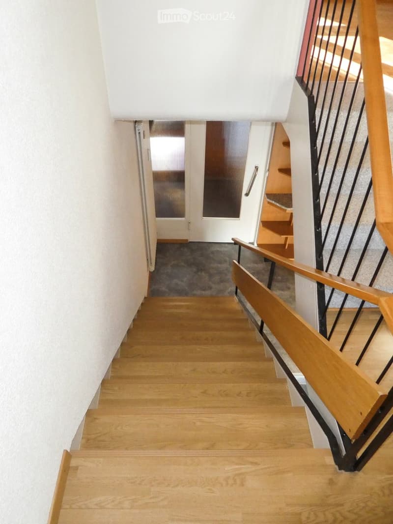 Wooden staircase with a wooden handrail and black metal balustrade, leading to a door at the end of the hallway.