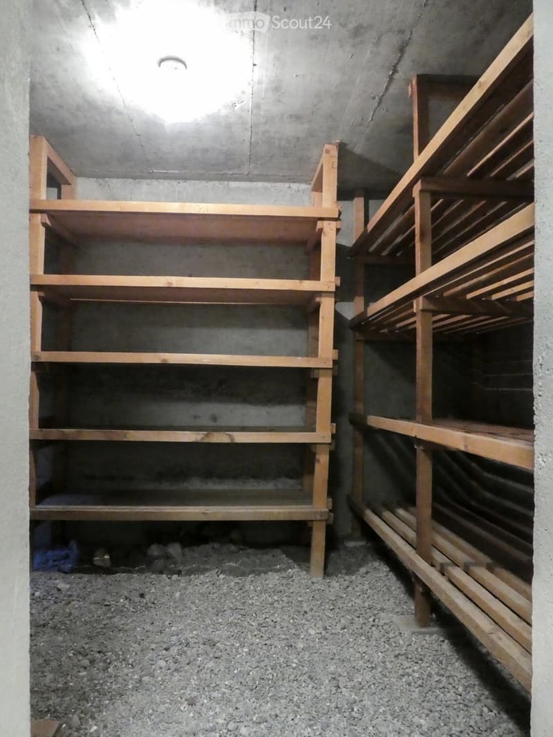 Empty storage room with concrete walls and wooden shelving units, gravel floor, and minimal lighting.