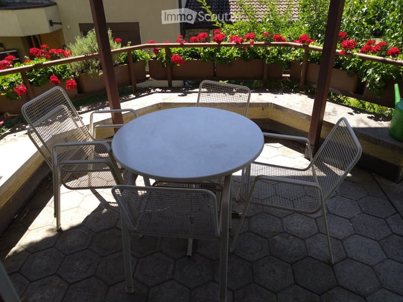 Balcony with a round table and four metal chairs, surrounded by flower boxes with red flowers.