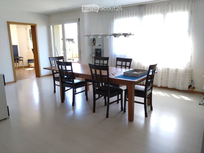 Bright dining room with a wooden dining table surrounded by six black chairs, large windows with white curtains, and a hanging light fixture above the table.