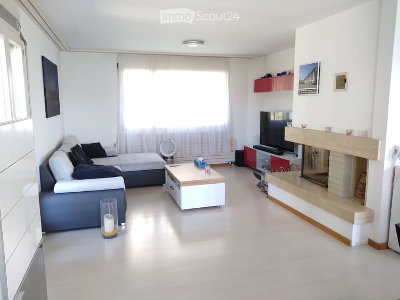 Modern living room with white walls, grey sofa, coffee table, fireplace, and TV. Features large windows with white curtains and a sleek design.