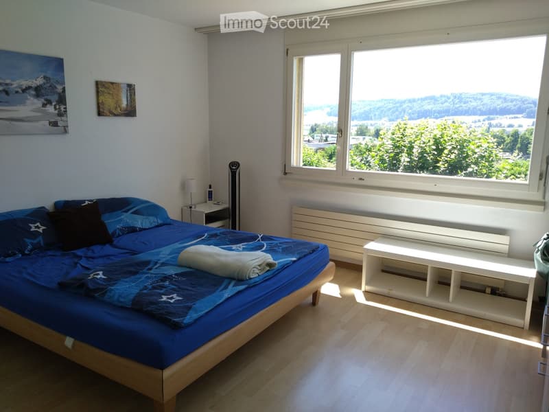 Bright bedroom with a double bed, nightstand, lamp, radiator, bench, and scenic view through large windows.