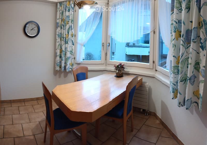 Dining room with a wooden table, four chairs, a flower pot on the table, and a clock on the wall. Large windows with floral curtains provide natural light.