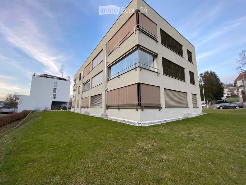 Modern apartment building with glass windows, beige exterior, and a grassy lawn in front.