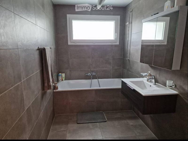 Modern bathroom with grey tiles, a bathtub, a sink with a mirror, and a towel hanging on the wall.