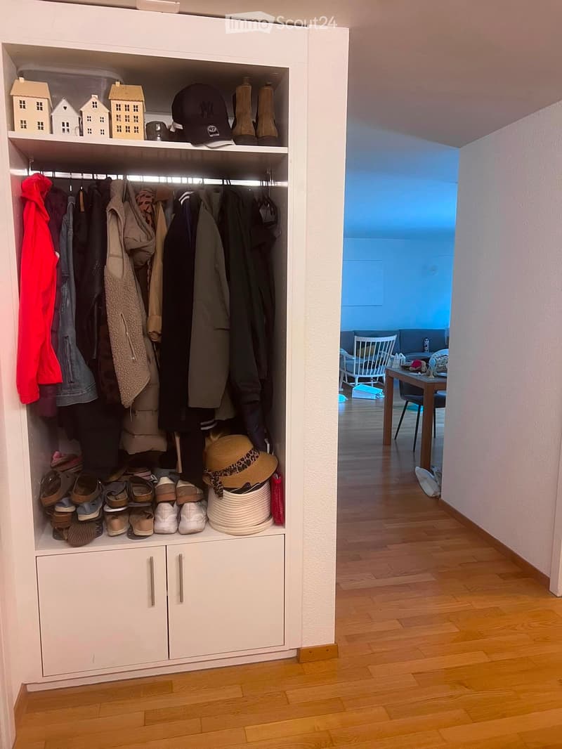A hallway with a built-in white closet containing clothing, shoes, and accessories. The hallway has wooden flooring and leads to a room with a table, chairs, and a white chair.