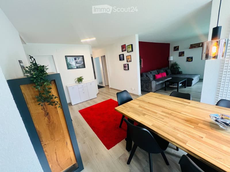 Modern open-plan living and dining area with wooden flooring, a red rug, a dining table with chairs, a sofa with cushions, a coffee table, and a white cabinet.