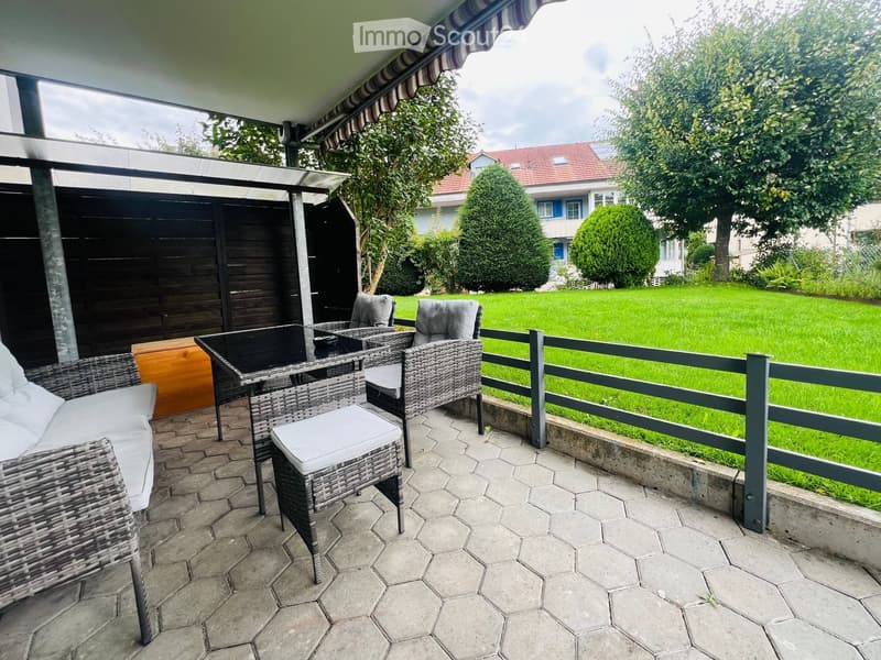 Outdoor patio with wicker chairs and a table, overlooking a well-maintained lawn and garden area.