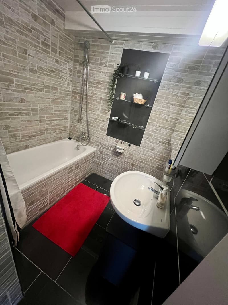 Modern bathroom with a white bathtub, a sink with a modern faucet, a mirror with shelves, and a red bath mat on the floor.