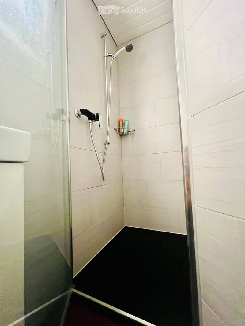 Modern shower area with white tiled walls, black floor, glass enclosure, and a handheld showerhead with a shelf holding bottles.