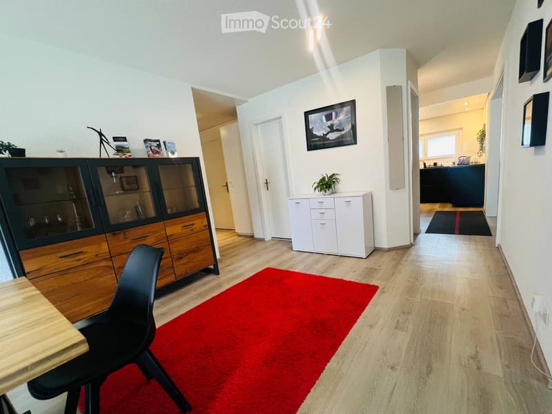 Modern living room with wooden flooring, red rug, black chair, small table, white cabinet, and a large display cabinet with glass doors.