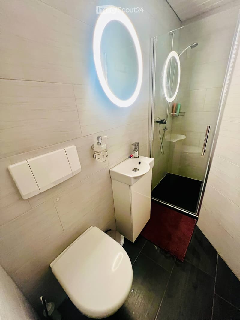 Modern bathroom with white tiles, a toilet, a sink with a modern faucet, a shower with a glass enclosure, and illuminated mirrors above the sink and shower.
