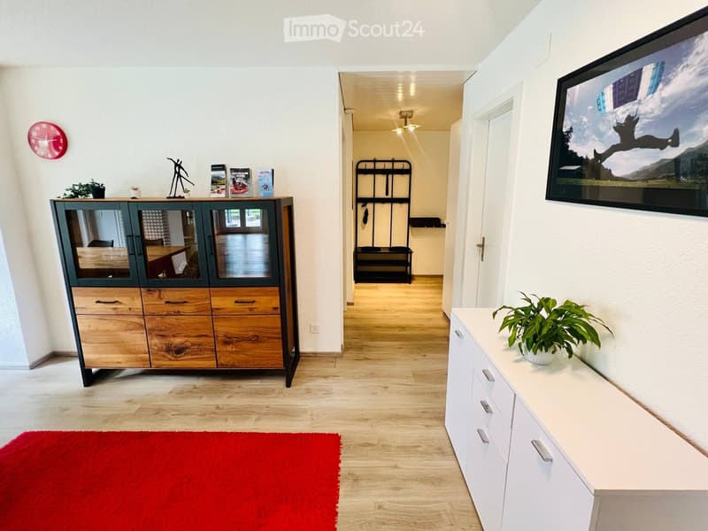 Bright hallway with wooden flooring, white walls, and a red rug. Features a wooden cabinet with glass doors, a white side table with a plant, a wall-mounted TV, and a coat rack in the background.