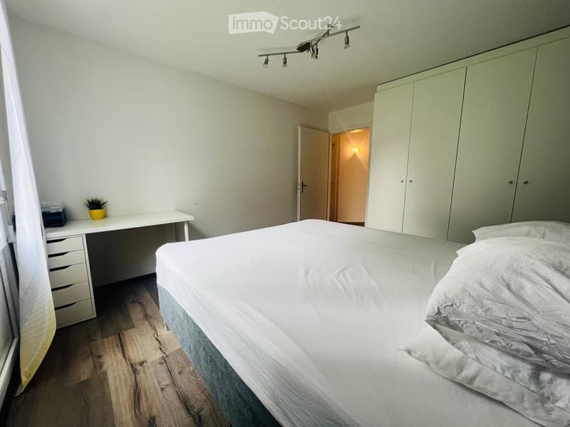 Modern bedroom with a large bed, white desk, small plant, and built-in wardrobe. Light wooden flooring and neutral-colored walls.