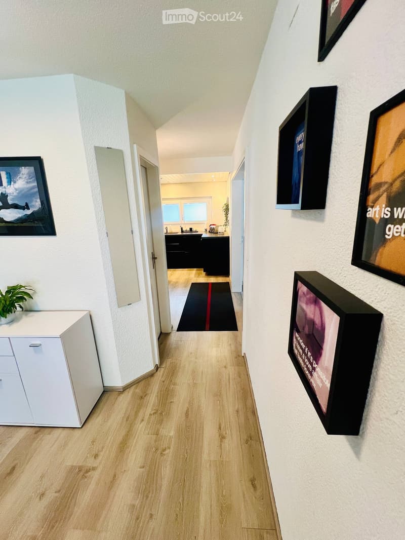 A modern hallway with light wooden flooring, white walls, and several framed artworks displayed on the right side. A small white cabinet with a potted plant sits on the left, and a black runner with a red stripe leads to an open doorway revealing a kitche