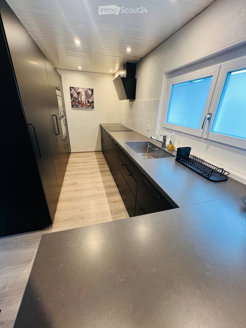 Modern kitchen with light-colored countertops, dark cabinets, stainless steel sink, and a window overlooking a blue sky.