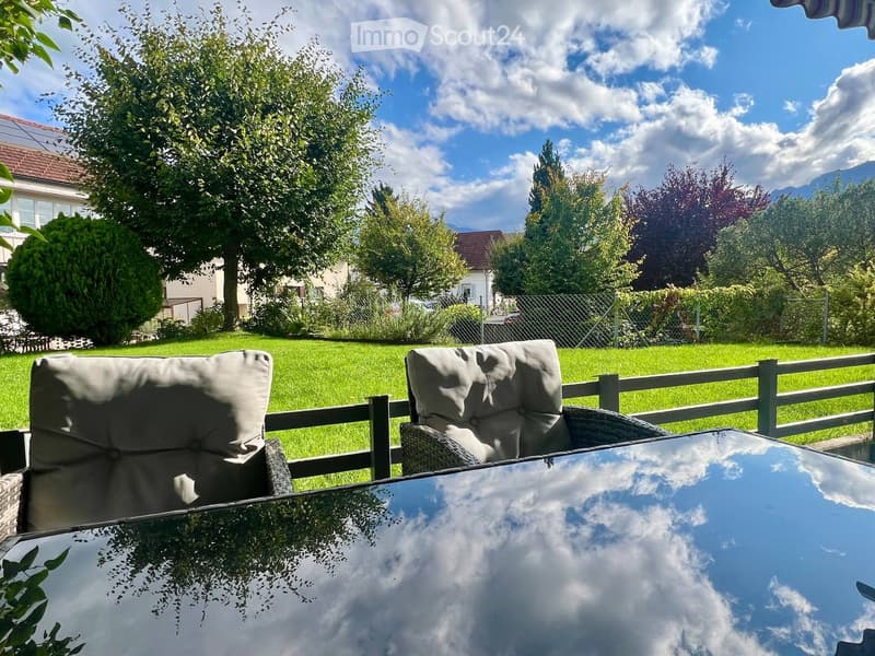 Outdoor patio with a glass table and two wicker chairs, surrounded by a wooden fence and a well-maintained lawn with trees and mountains in the background.