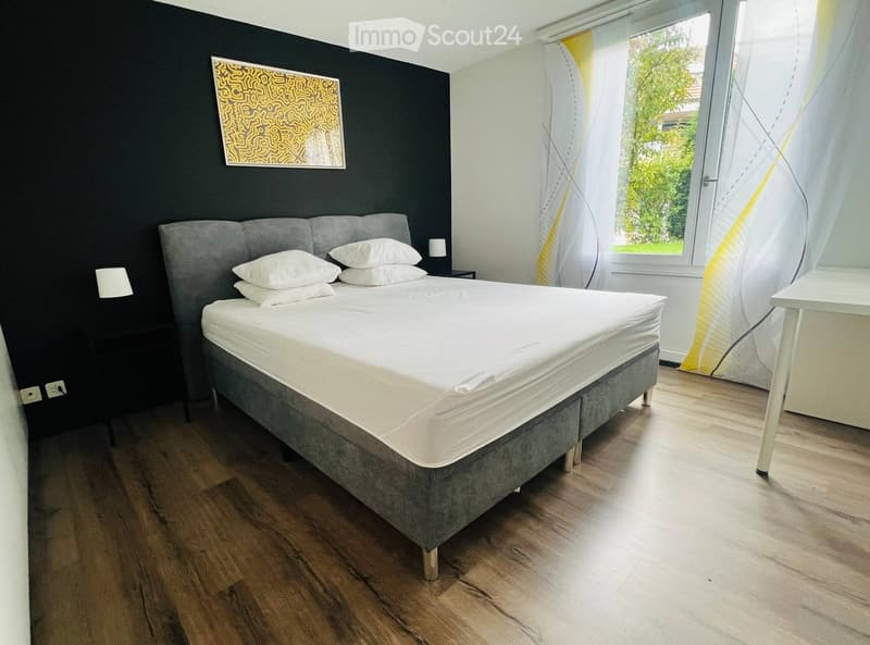 Modern bedroom with a large bed, nightstand, lamp, and a small table near the window. The room features wooden flooring and large windows with sheer curtains.