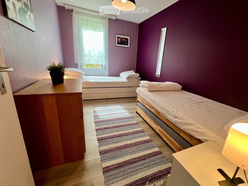 Cozy bedroom with two single beds, nightstands, a lamp, a colorful rug, a small plant, and a mirror on the wall.