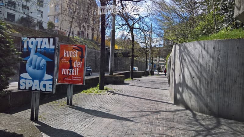 A paved outdoor area with posters, trees, and a raised ground floor visible in the background.