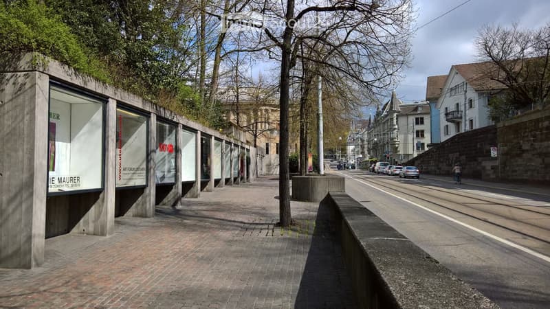 Urban corridor with brick pavement, trees, and a tram line running alongside, featuring modern concrete structures and signage.