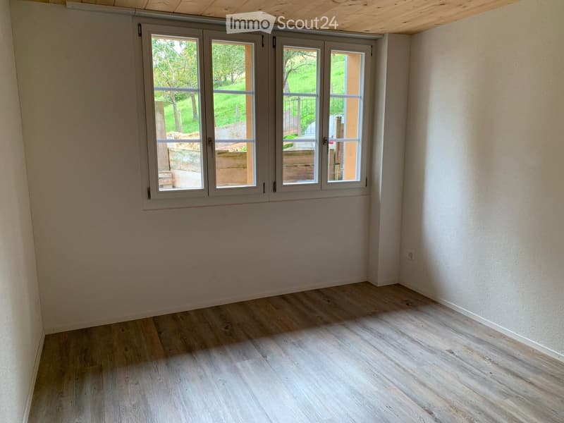 Empty room with light wooden flooring, white walls, and large windows offering a view of a green outdoor area.