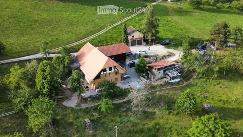 Aerial view of a two-story house with a garage, surrounded by greenery and a parking area.