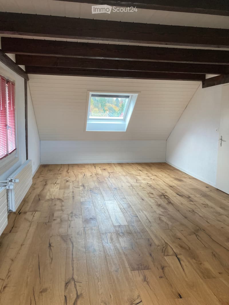 Empty attic room with wooden floors, white walls, exposed wooden beams, and a skylight window.