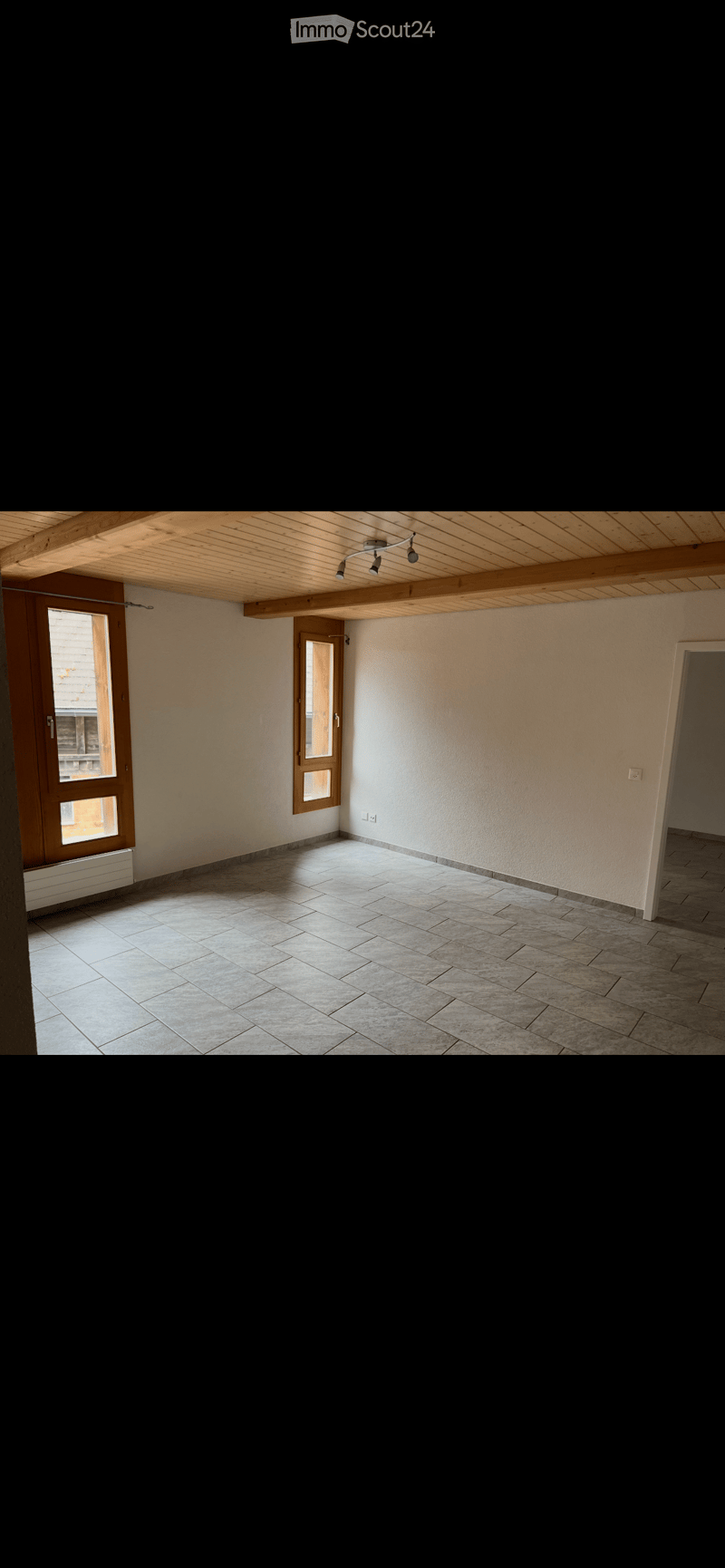 Empty room with tiled floor, wooden ceiling beams, and two windows with wooden frames. No furniture or equipment present.