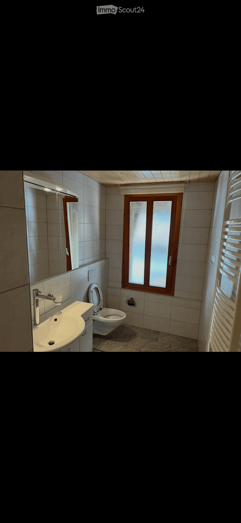 Modern bathroom with a white sink, chrome faucet, toilet, mirror, and a radiator. The room features tiled walls and a window with frosted glass.